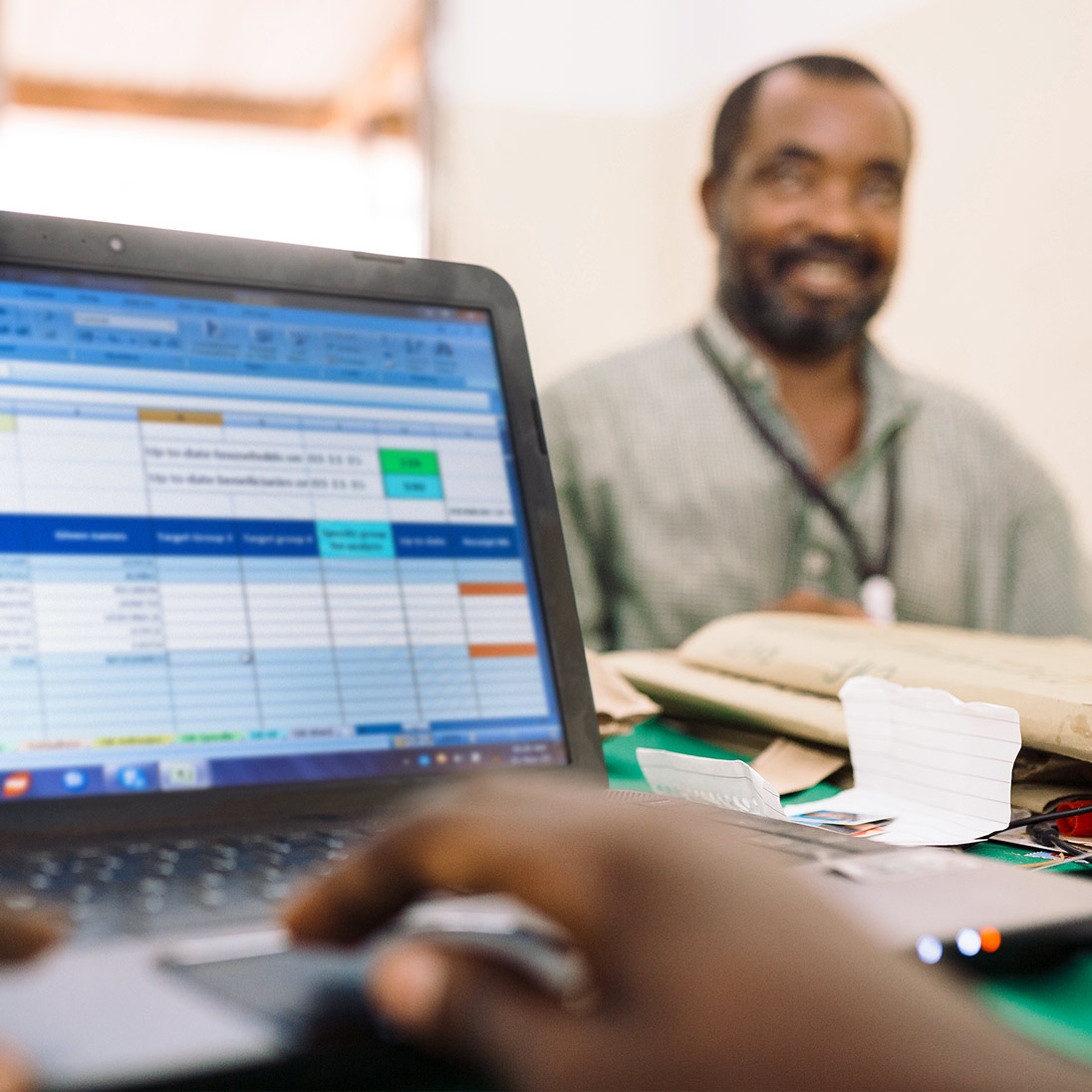 A person typing on a laptop showing an Excel table; in the background, a man sits at a desk smiling.
