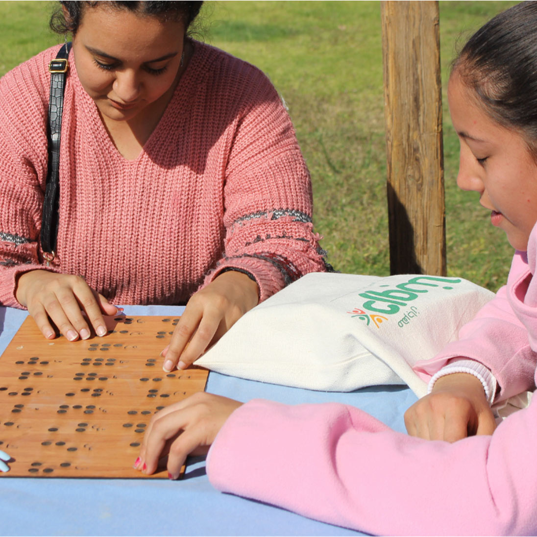 Three young women sitting at a table outdoors run their fingers over a wooden board with Braille.