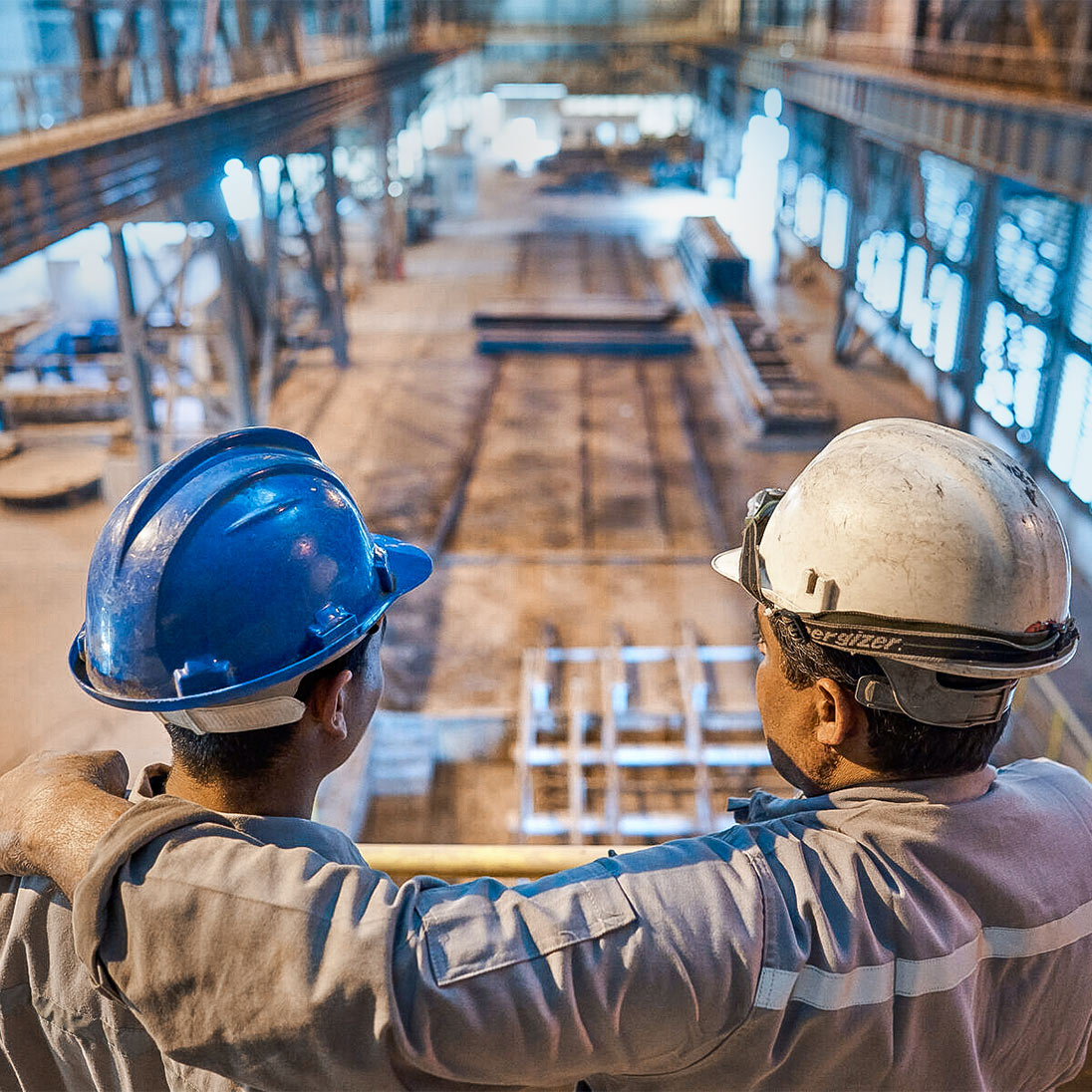 Two men in hard hats stand on a gallery looking down on a large industrial hall with machines and work areas.